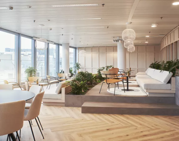 Spaceous and illuminated lounge area with large windows, round tables, chairs plants and a sofa at Stibbe in Brussels