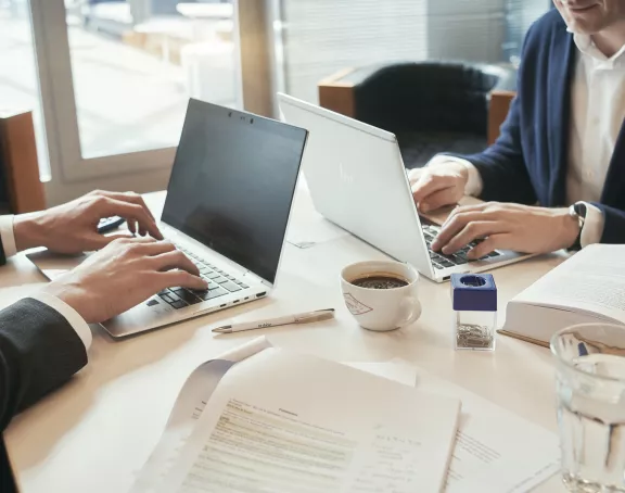 Cropped close-up of two business people sitting opposite each other at a conference table and working on their laptops