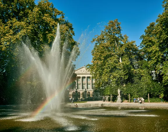 Brussels park scene featuring a fountain with a rainbow, vibrant green trees, and the Royal Palace of Brussels in the background
