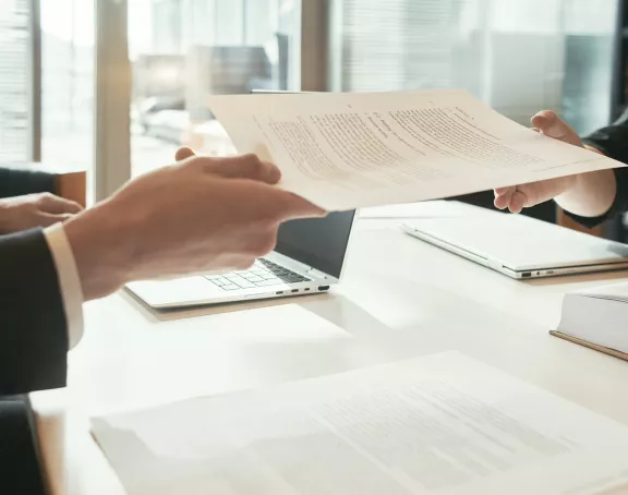 Cropped close-up of two business people sitting opposite each other at a conference table with one of the two handing over a document to the other person