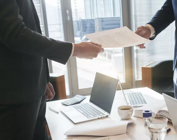 Cropped close-up of two business people standing around a conference table with one of the two handing over a document to the other person