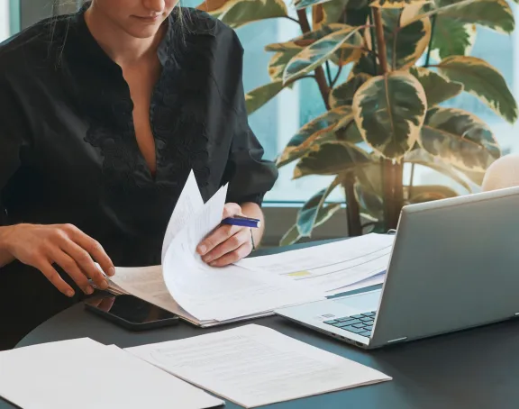 Cropped close-up of a woman sitting at a desk and leafing through some papers with a laptop in front of her
