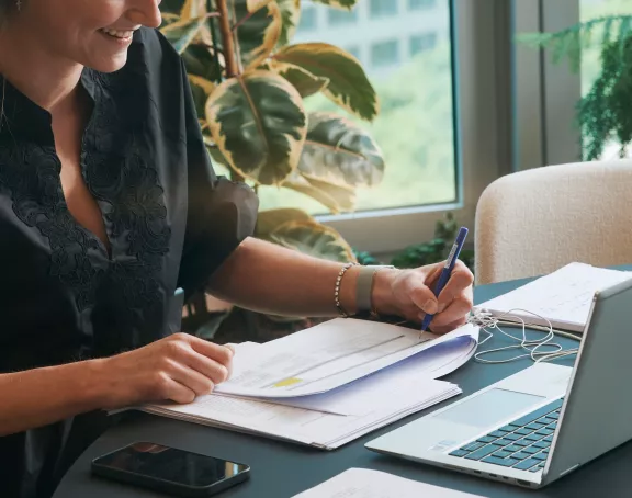 Cropped close-up of a woman sitting at a desk and taking notes on a document with a laptop in front of her