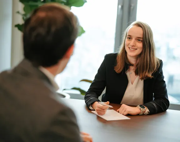 A female Stibbe lawyer sitting at a table in a meeting room, smiling while holding a pen and notepad. She is looking toward her conversation partner across the table, who is facing the camera with his back.