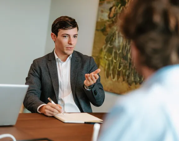 A male Stibbe lawyer seated at a table in a meeting room, gesturing with his hands while speaking to his conversation partner across the table, whose back is turned to the camera.