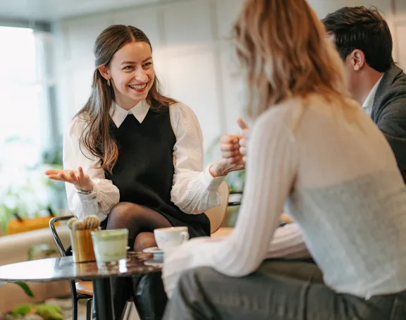 Two women and one man, all co-workers, engaging in a friendly conversation. The focus is on one woman, smiling broadly, while the other two are facing their backs to the camera, listening and smiling.