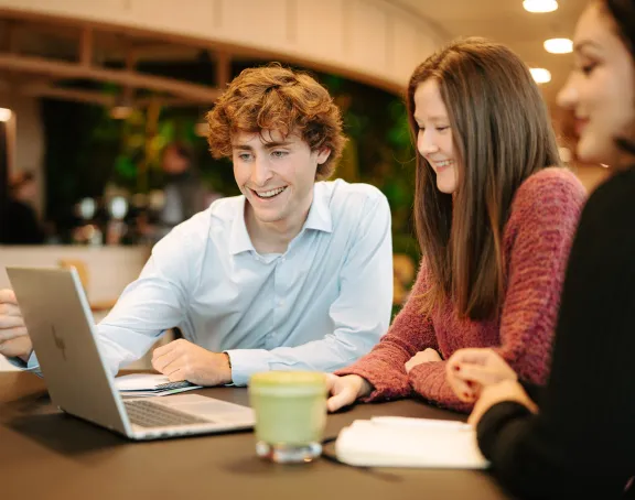 Two female Stibbe employees and one male Stibbe lawyer sitting together at a table. The man is smiling and pointing at a laptop screen, while the women are looking at the screen with interest. The group looks engaged and happy.