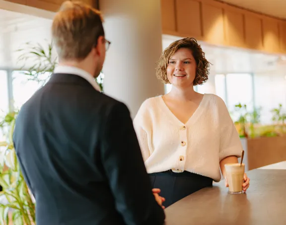 A woman and a man having a conversation at the counter of the coffee bar at Stibbe in Brussels. The man is facing away from the camera, while the woman smiles and looks at him, holding a cup of coffee in her hand.