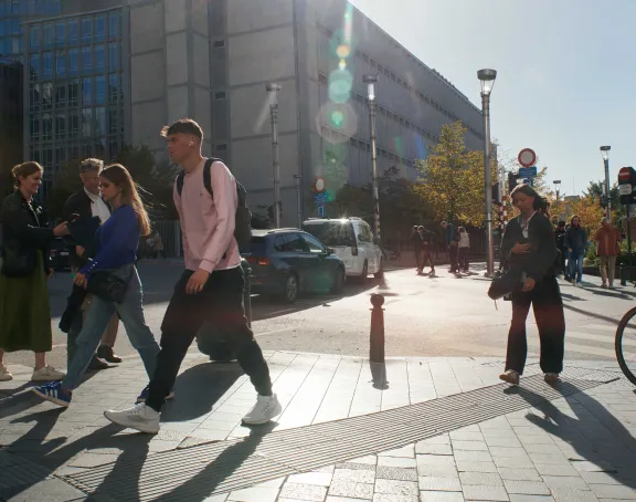 Pedestrians cross a sunny street in Brussels with modern and historic buildings and autumn trees in the background.