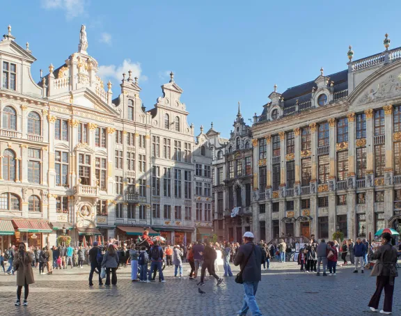 A wide view of the Grand Place in Brussels, showing the bustling square under a bright blue sky. The square is filled with people walking and observing street art displays, surrounded by richly decorated historical buildings.