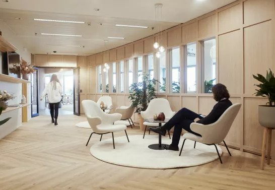 Corridor to the lounge space at Stibbe in Brussels featuring a man sitting in a chair looking at his phone and a woman walking down the corridor about to enter the lounge space