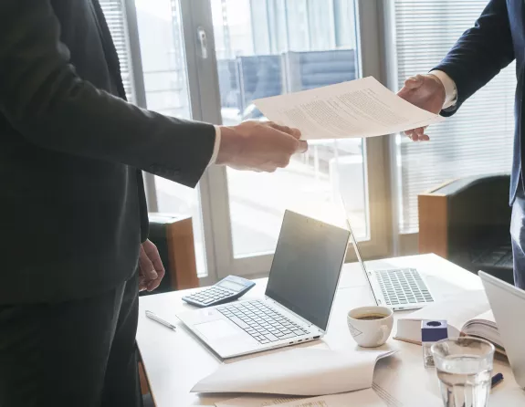 Cropped close-up of two business people standing around a conference table with one of the two handing over a document to the other person