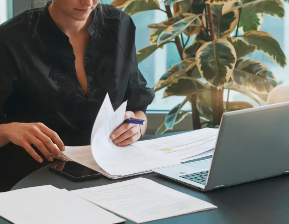 Cropped close-up of a woman sitting at a desk and leafing through some papers with a laptop in front of her