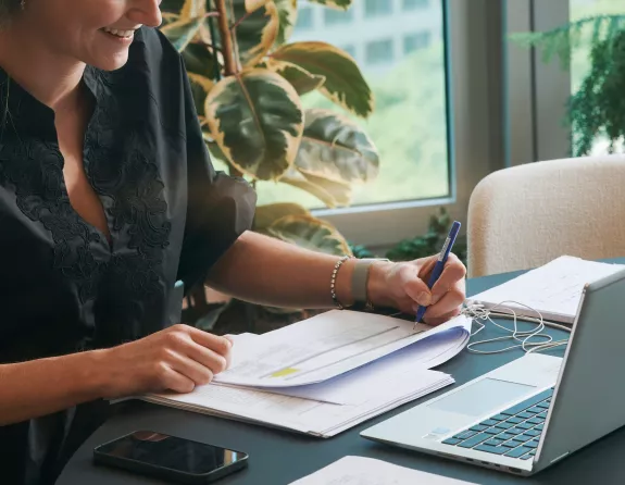 Cropped close-up of a woman sitting at a desk and taking notes on a document with a laptop in front of her