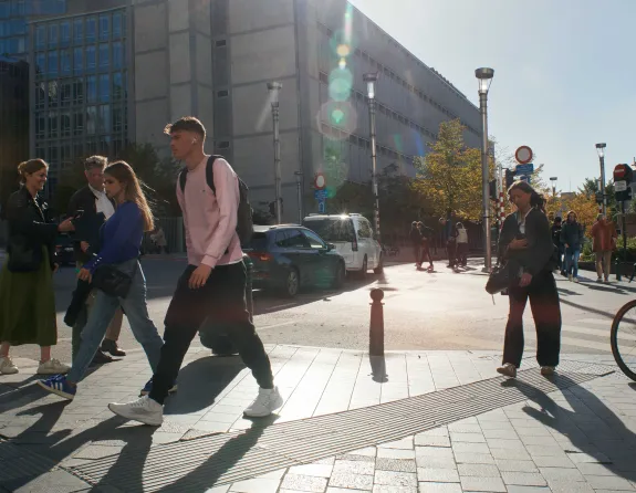 Pedestrians cross a sunny street in Brussels with modern and historic buildings and autumn trees in the background.
