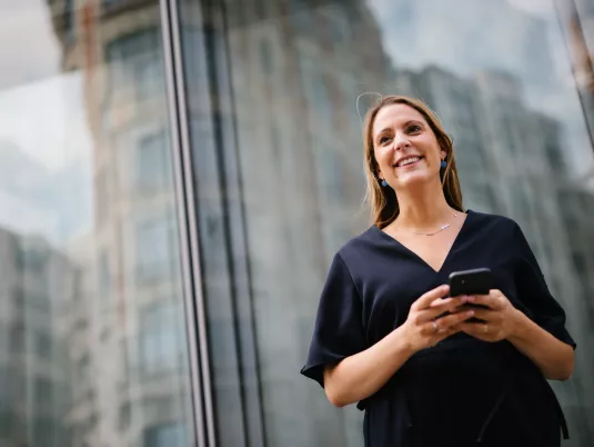 Female Stibbe lawyer standing in front of the entrance to the Central Plaza building in Brussels, smiling and looking ahead whilde holding her smartphone with both hands