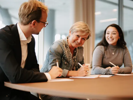 Three co-workers, two women and one man, sitting side by side at a table, laughing together. The co-worker in the middle is looking at the camera and smiling, while the others are facing each other.