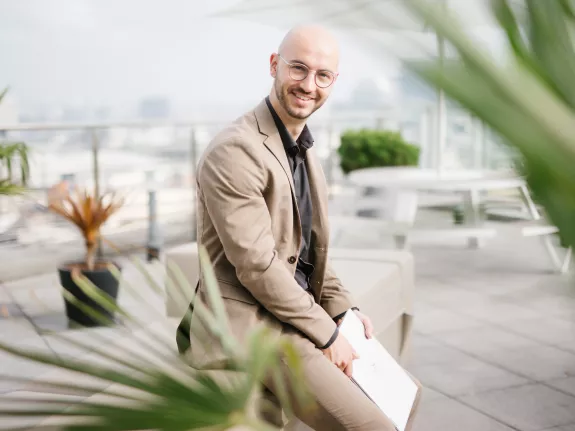 Male Stibbe lawyer smiling at the camera while leaning against a sofa and holding his laptop on the rooftop terrace of Stibbe in Brussels