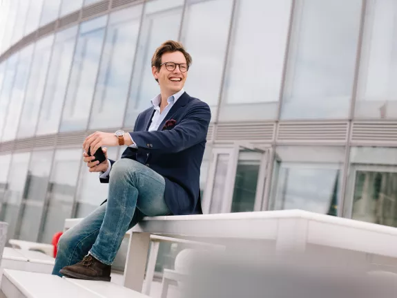 Male Stibbe lawyer sitting on a table with one foot resting on the table bench on the rooftop terrace at Stibbe in Brussels, looking sideways and smiling while holding his smartphone in both hands