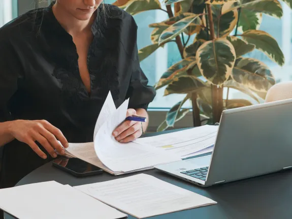 Cropped close-up of a woman sitting at a desk and leafing through some papers with a laptop in front of her